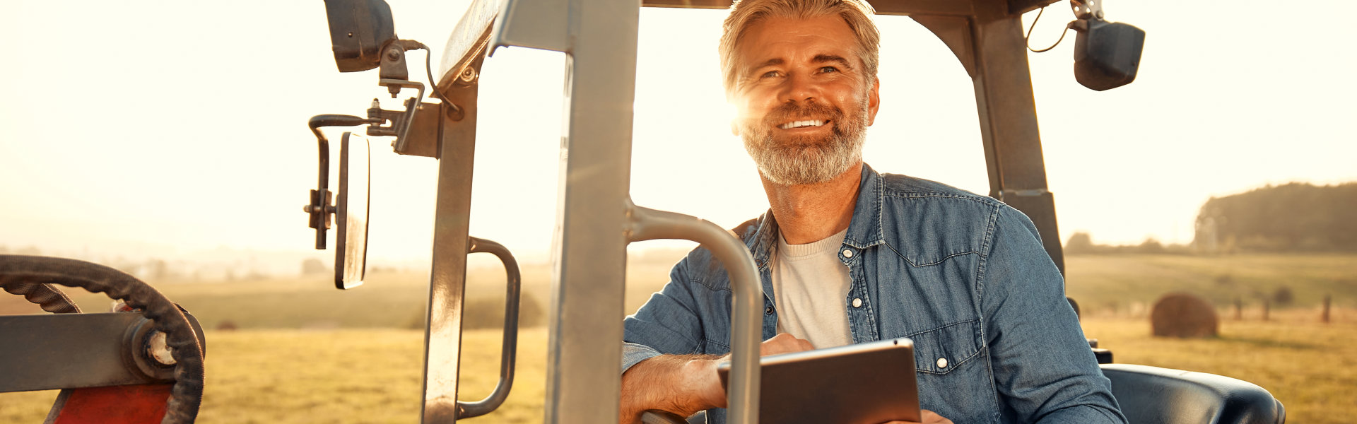 Smiling man in a denim shirt sits inside a tractor, holding a tablet. Sunlight fills the rural field background, creating a warm, optimistic tone.